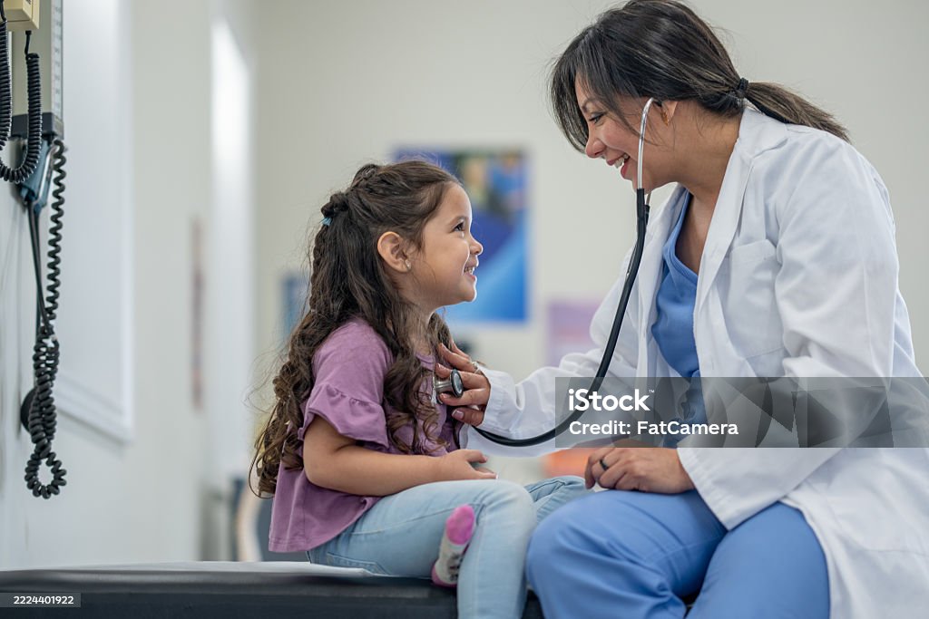 A sweet little girl sits up on an exam table during a doctors appointment. She is dressed casually and her female doctor is seated in front of her as she listens to her heart.