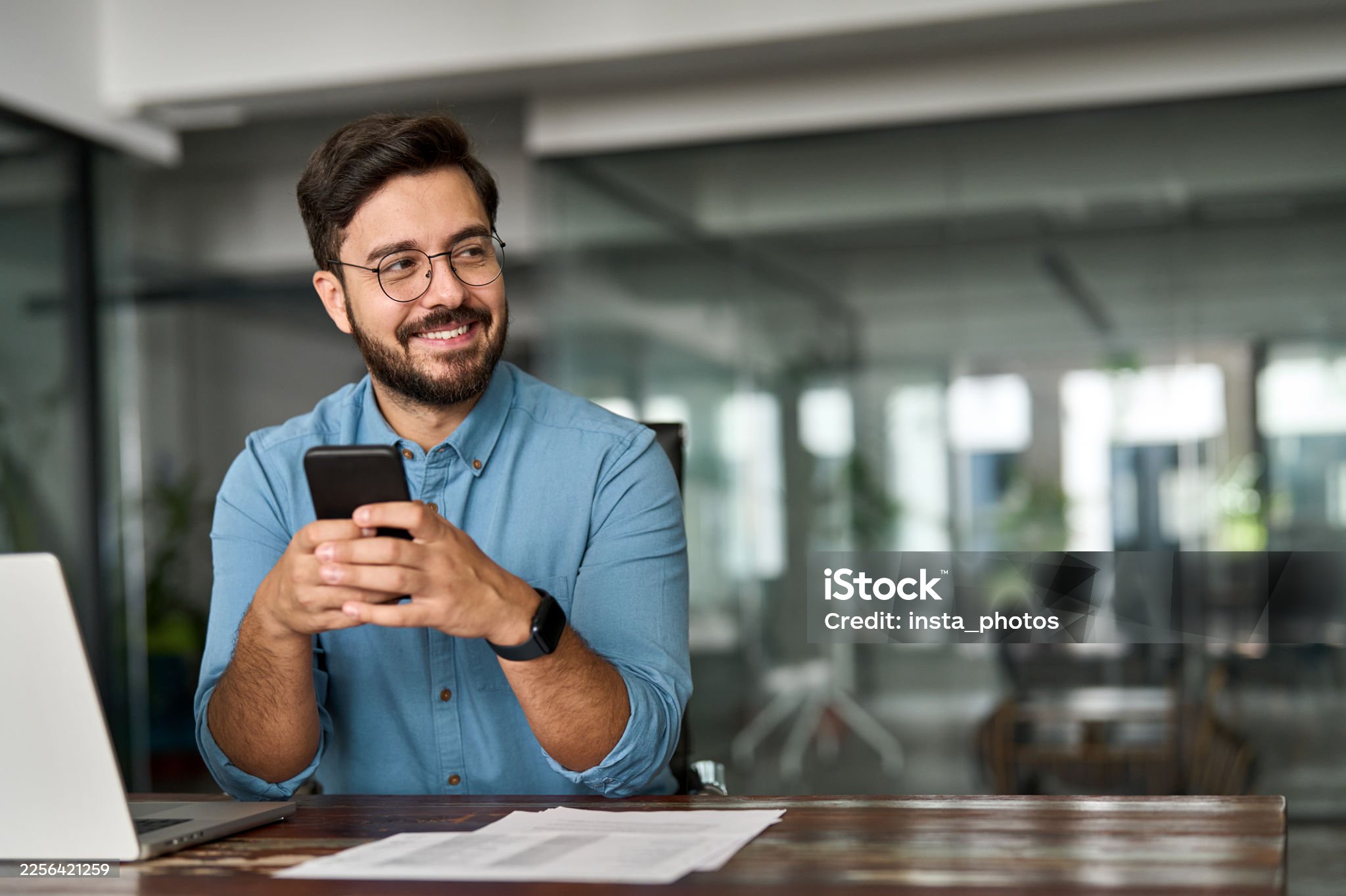 Busy happy young Latin business man holding smartphone sitting in office. Professional hispanic businessman entrepreneur or manager using financial banking apps on cell phone technology at work.