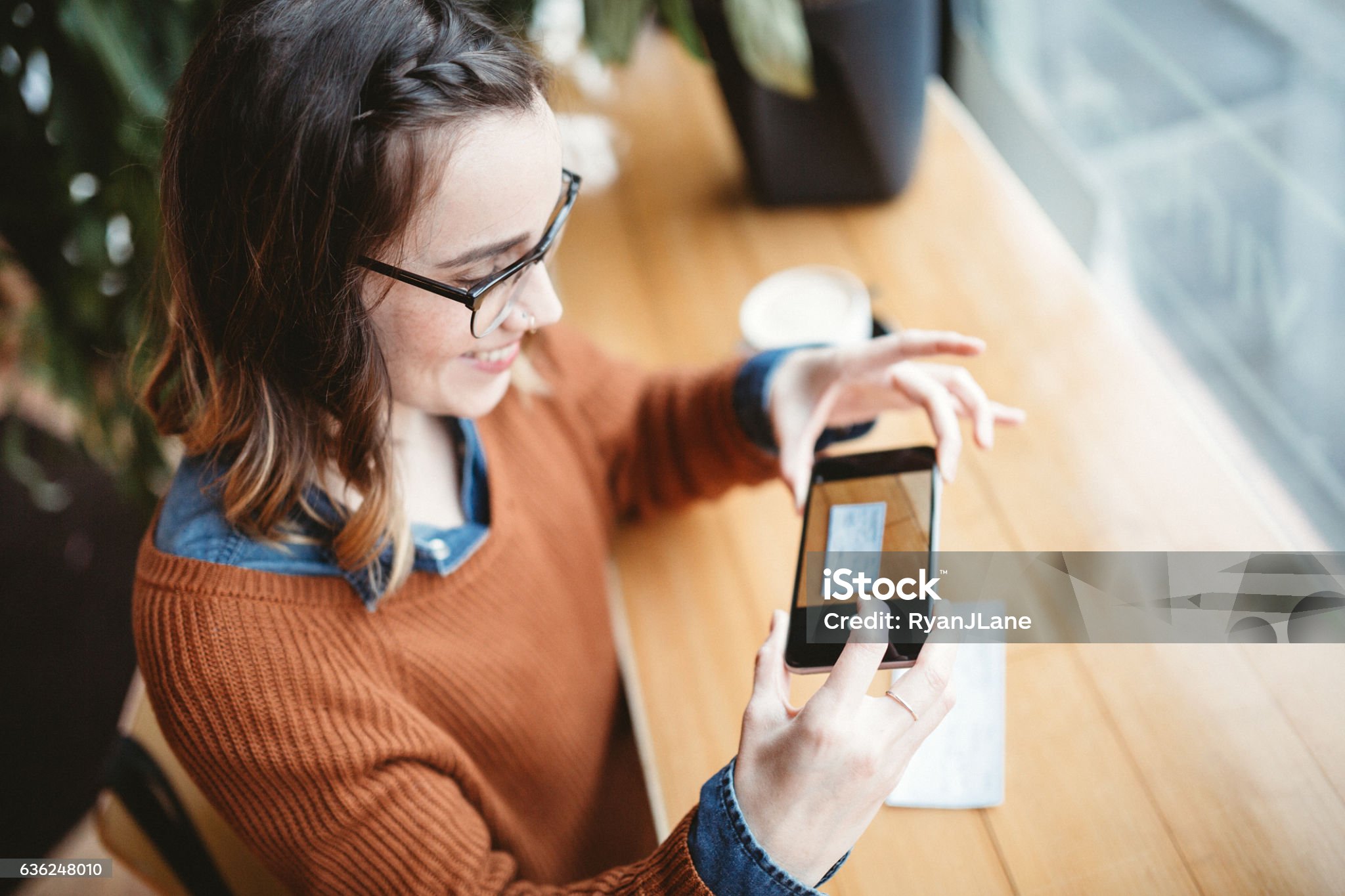 A smiling young woman takes a picture with her smart phone of a check or paycheck for digital electronic depositing, also known as "Remote Deposit Capture". She sits in a coffee shop, enjoying an espresso latte. Bright sunlight shines in the window. Horizontal image.