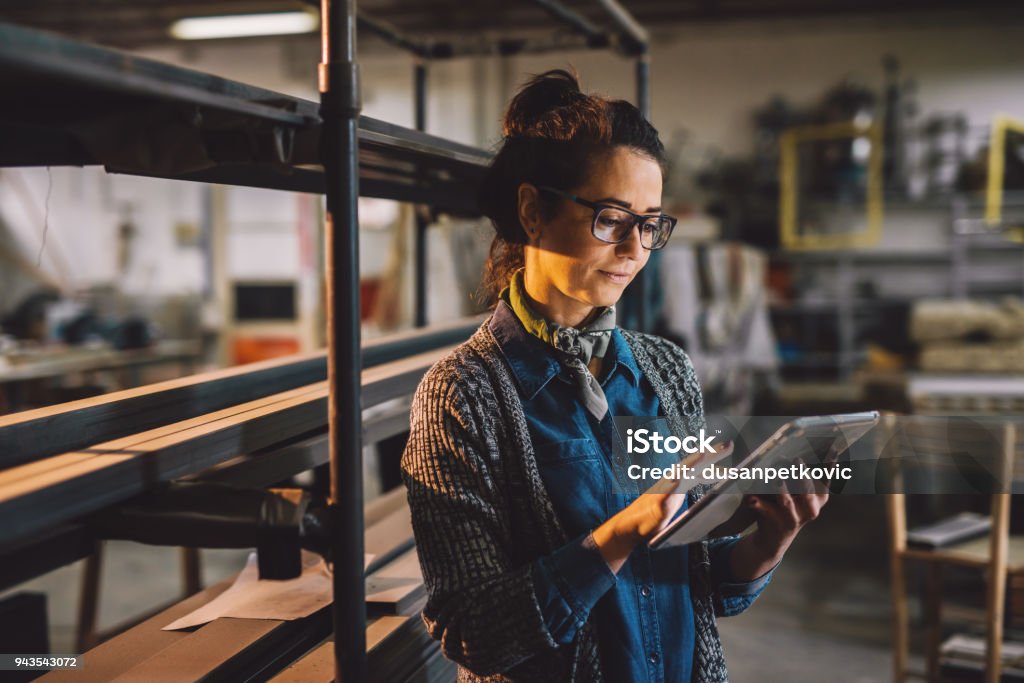 Close up view of hardworking focused professional motivated business woman holding a tablet next to the shelf with metal pipes in the fabric workshop.